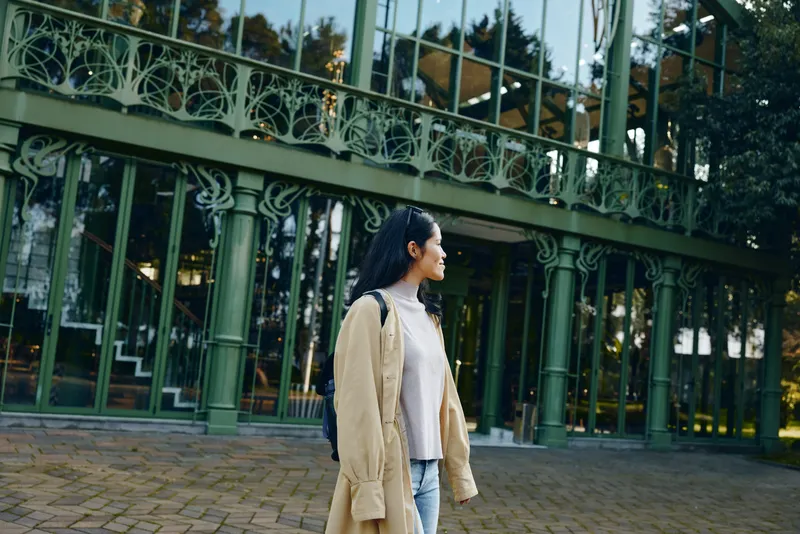 Woman standing outdoors in a beige coat beside a green iron railing and glass building, casual style, calm street scene, urban lifestyle photography
