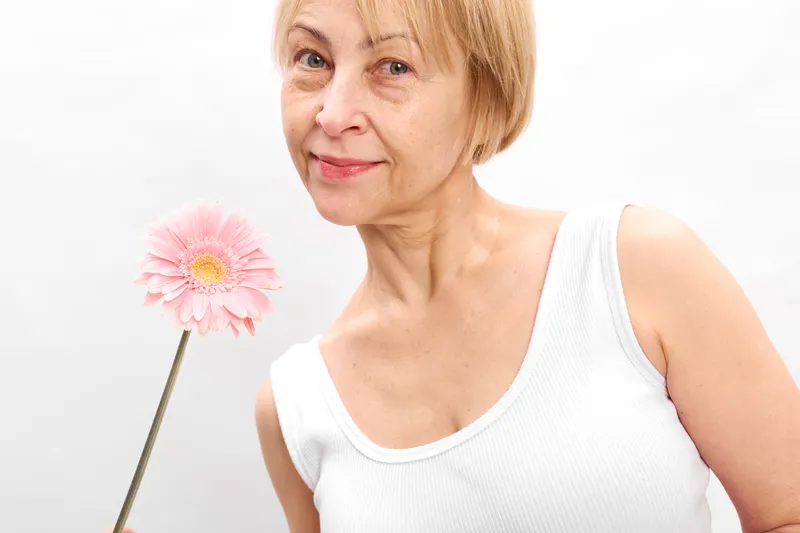 Elderly woman portrait in a bright studio, holding a pink flower on white background, gentle smile and calm pose. Concept of spring beauty, wellness, skincare for mature age.