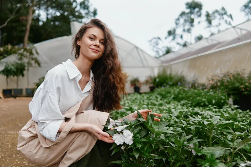 Young woman in a garden showing flowers, wearing a casual white shirt and embracing nature, surrounded by greenery and vibrant plants, reflecting joy and serenity