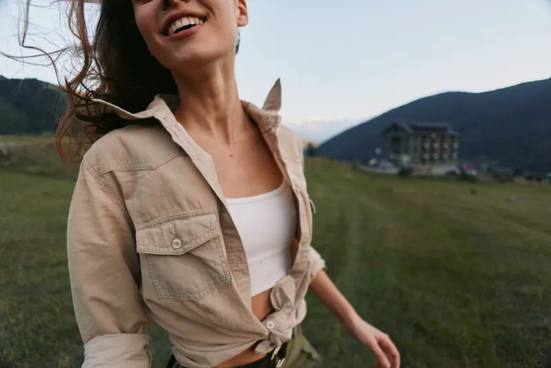 Happy woman in beige shirt and white top walking outdoors in a green field with mountains in the background. Concept of lifestyle, freedom, and natural beauty.
