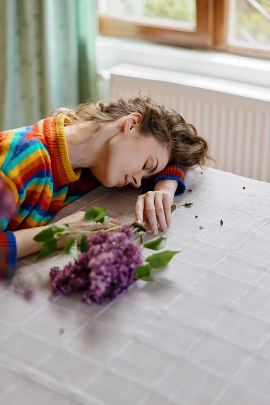 Woman resting at a home table, sleeping with her head on her arm, colorful sweater, soft natural light, fresh flowers nearby, peaceful mood, intimate everyday moment indoors