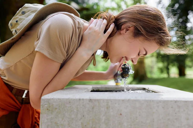 Young woman drinking water from a public fountain in a park, leaning over the stone basin with a backpack, casual summer outfit and smile while refreshing and hydrating outdoors.