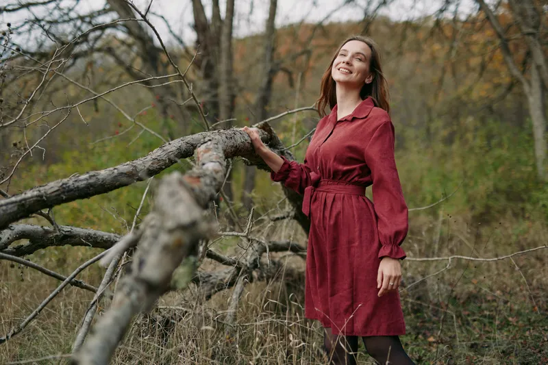 Smiling woman in a red dress stands among fallen branches in a quiet forest clearing, conveying natural warmth and authenticity through candid posture and genuine expression, capturing an unposed