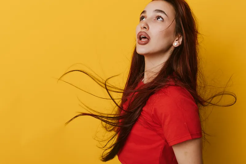Young woman with long hair wearing red shirt looking up with surprised expression on yellow background. Portrait of female model with flowing hair and open mouth in studio.