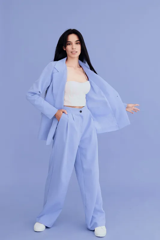 woman in pastel blue suit posing confidently, wearing white crop top and sneakers against solid lavender background, stylish young adult with long dark hair.
