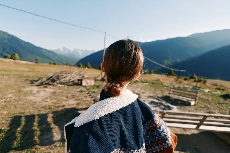woman back in jacket with tied hair and shearling collar standing in a meadow by a bench and swing, overlooking mountains and countryside landscape under warm sunlight and open horizon