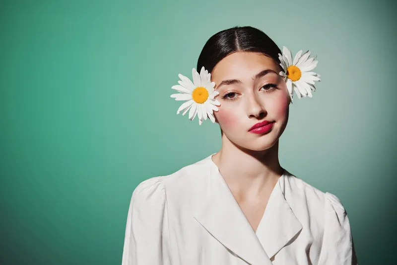 Young woman with flowers in hair poses in white blazer. Soft makeup and sleek hairstyle on green background. Creative beauty fashion editorial concept portrait.