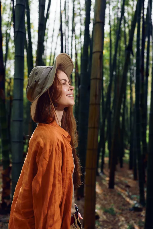 Woman bamboo forest hat smile nature outdoors portrait in orange jacket, side profile among tall trunks and green foliage, serene travel scene, peaceful expression and soft natural light.