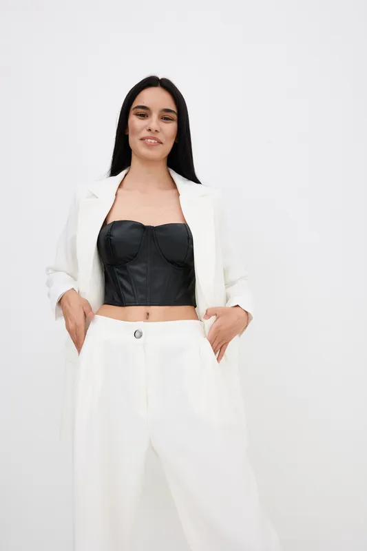 Smiling woman in white suit and black bustier stands confidently with hands in pockets against plain white background, showcasing modern fashion and relaxed pose.