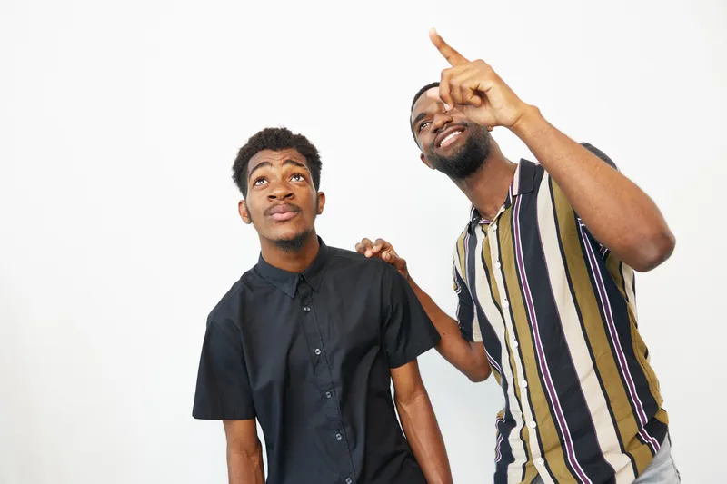 Two joyful Black men express enthusiasm while pointing upwards, showcasing their friendship and excitement in a bright, contemporary setting filled with positivity.