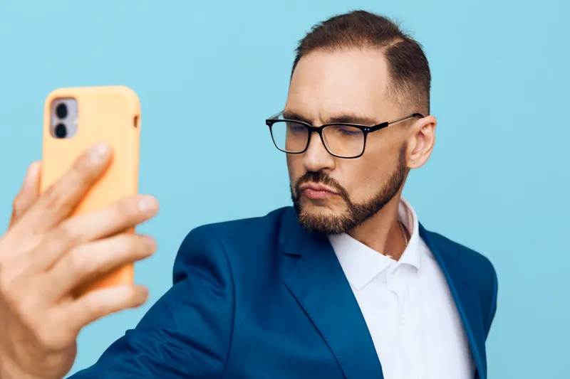 A focused business man in a blue suit takes a selfie against a solid colored background, showing confidence, concentration, and subtle emotion in a modern work moment.