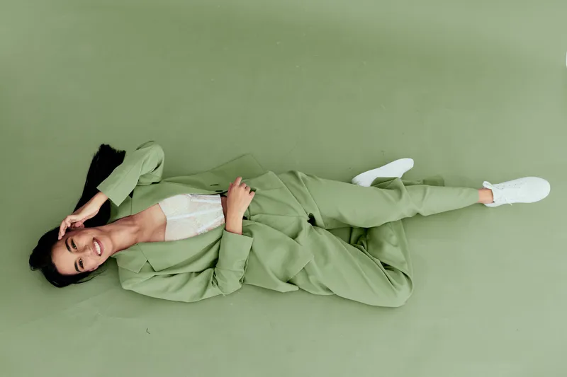 Young woman in green suit lying on floor smiling brightly, wearing white sneakers and lace top, casual stylish outfit with fresh and relaxed mood.