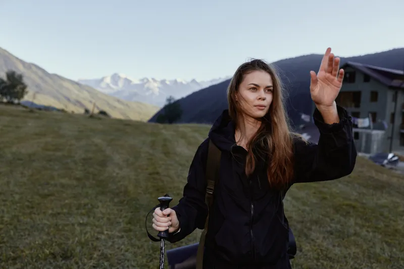 A female hiker stands outdoors in a grassy alpine landscape, holding a trekking pole and looking ahead with a determined expression, ready for an adventurous mountain journey.