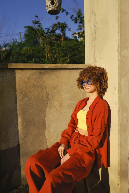 A stylish woman sits on a stone ledge in a warm orange suit and yellow top, sunglasses on, enjoying sunny outdoor light. Concept for casual fashion and outdoor lifestyle