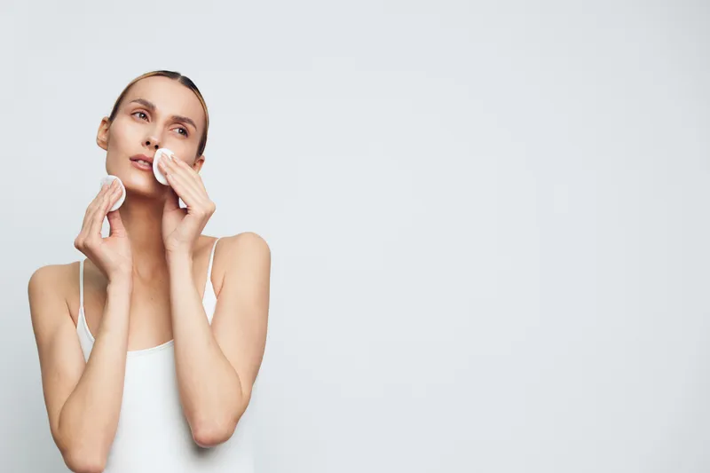 Smiling woman with clear skin applying cotton pads to cleanse her face in a bright studio. Focus on skincare, self care, and gentle cleansing with soft lighting.