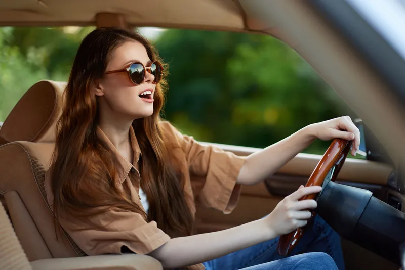 Cheerful young woman driving a vintage car, wearing sunglasses, enjoying a sunny day with a lush green background, perfect for travel and adventure themes.