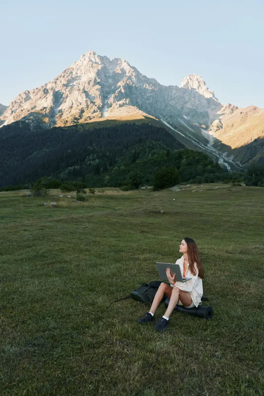 A young woman sits on a grassy field with a laptop, outdoors beneath towering rocky peaks. She works calmly, focused and content, amid a serene natural setting.