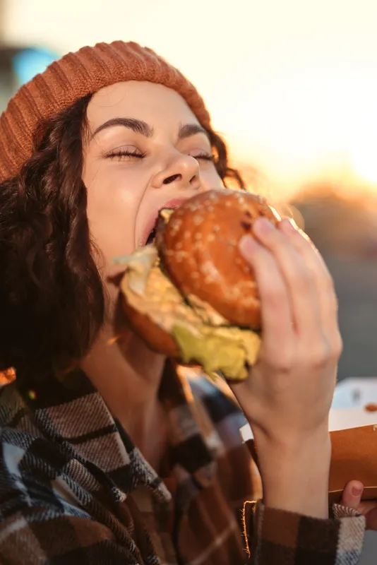 woman burger eating outdoor candid smile beanie plaid enjoying a big bite at golden hour glow, authenticity and candid lifestyle with emotional storytelling and mindful living.