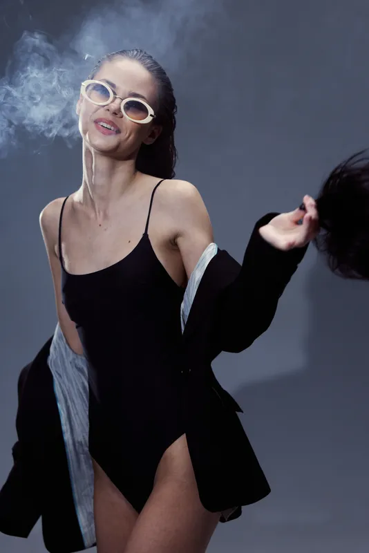 Smiling woman in black swimsuit and jacket wearing sunglasses with smoke against dark background.