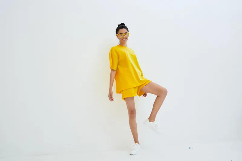 young woman in bright yellow casual outfit posing confidently against white background, wearing sneakers and glasses, radiating joy and energy