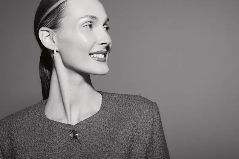 Smiling Caucasian woman with light skin and pulled back hair wears textured top and earrings, looking right with bright studio lighting and gray background.
