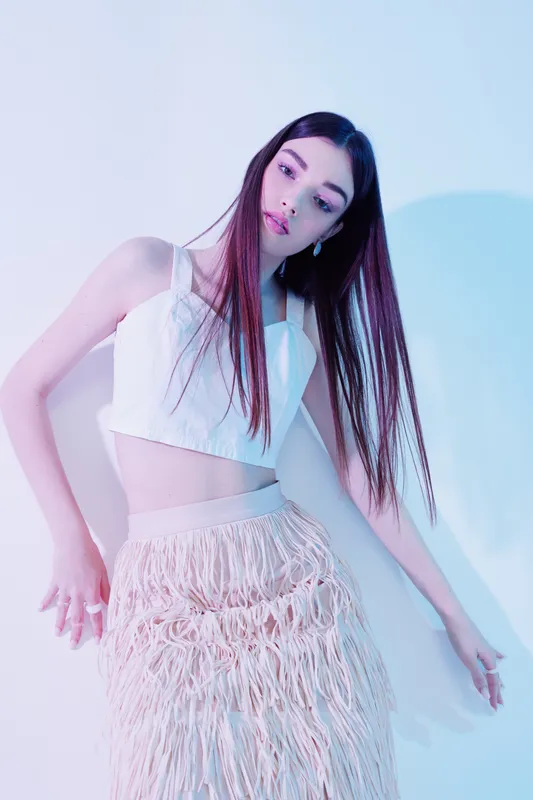 Young woman with long straight hair wearing a white crop top and textured skirt posing in soft pastel lighting. Concept for woman beauty fashion and modern style.