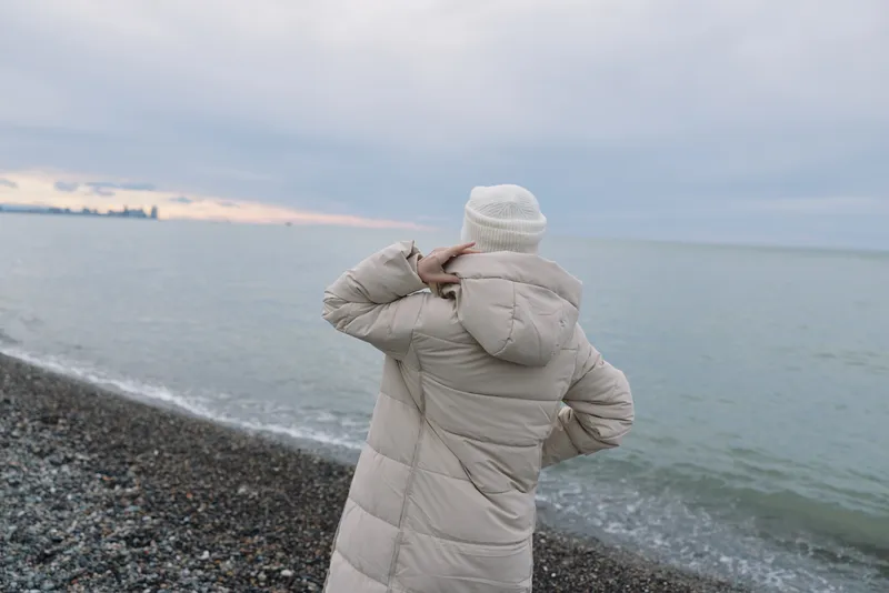 Beach seascape in winter: a person in a coat and hat stands on pebbles, gazing at the calm water and distant horizon, creating a quiet mood.
