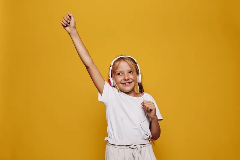 Smiling girl in white t-shirt wearing headphones dances with raised arm on yellow background. Happy child enjoying music and expressing excitement. Studio shot with vibrant colors.