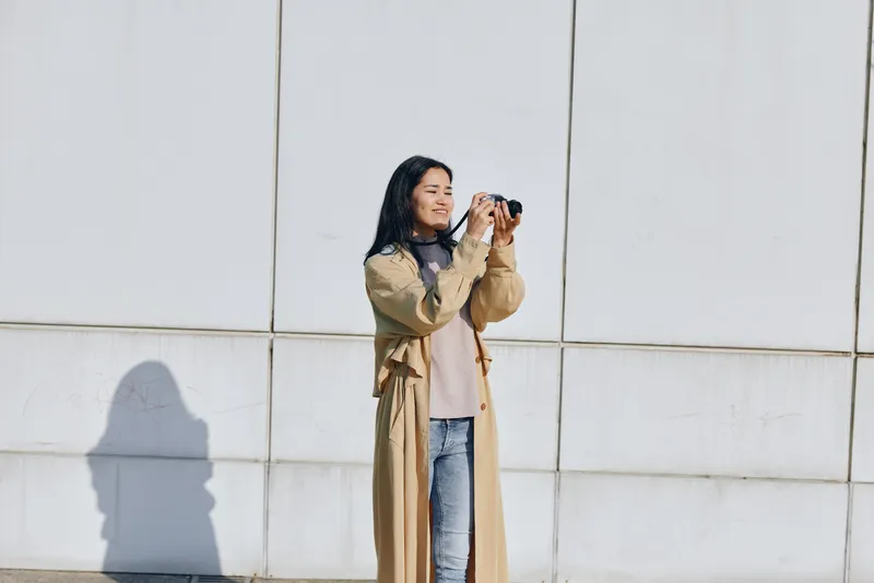 Woman with camera outdoors, capturing light tiled wall backdrop in a candid street photography moment, wearing a long beige coat and jeans, showcasing style and curiosity in urban exploration