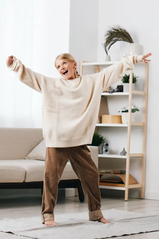 Playful child in a cozy oversized sweater dances with arms wide, filling a bright living room with warmth, movement, and carefree cheer during a sunny day indoors.