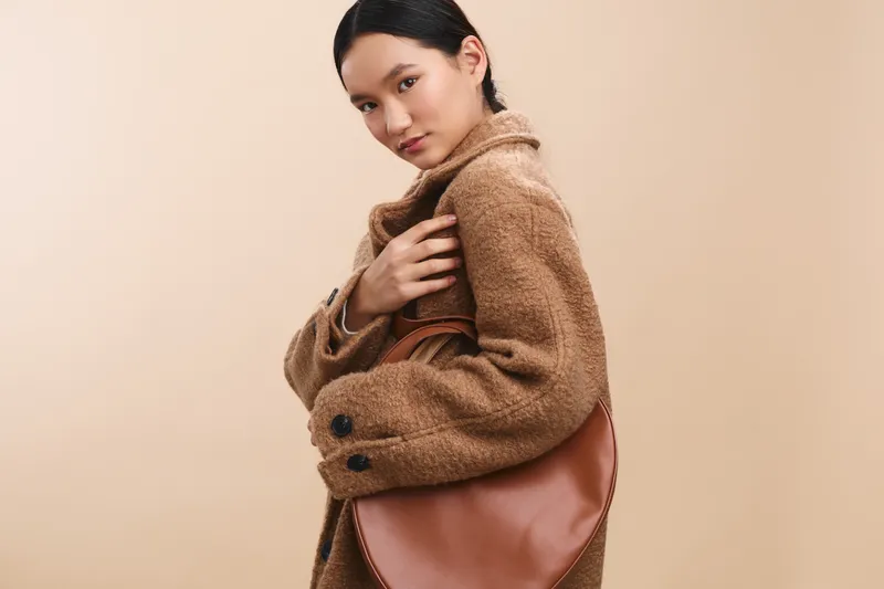 Asian woman wearing a cozy brown coat carries a leather bag, posing against a warm neutral backdrop to convey calm confidence and subtle elegance in a studio setting.
