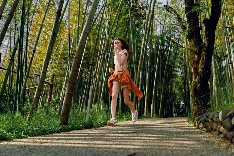 Woman running along a bamboo forest path, smiling and jogging with smartphone and headphones. Casual outfit with shorts and sweater tied around waist during outdoor fitness session.