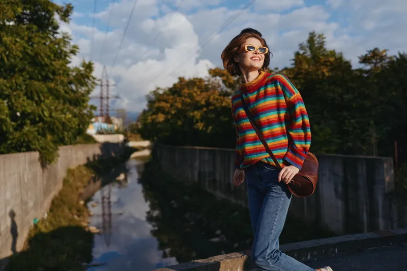 Woman in a rainbow sweater with sunglasses and shoulder bag standing by a canal smiling, wearing jeans in urban park scene with trees and blue sky for casual outdoor lifestyle.