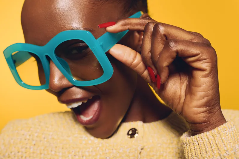 Close-up of a joyful black woman with dark skin wearing large turquoise glasses and a beige sweater. She smiles with red nails and a yellow background. Concept of fashion and happiness.