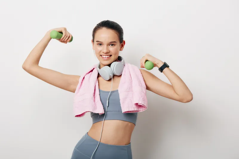 Fitness woman in workout mode lifting light dumbbells with a towel draped over shoulders and headphones, exuding energy and strength for a healthy routine.