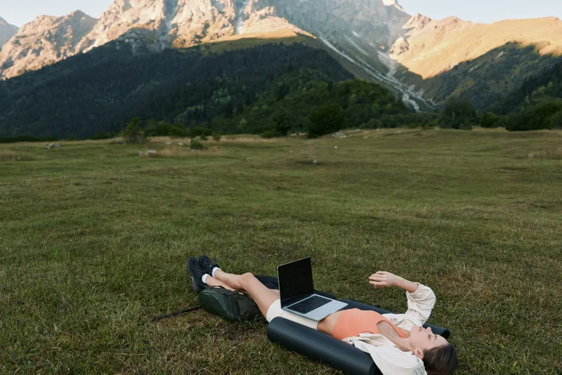 An outdoor work scene featuring a person lying on a grassy field with a laptop, set against distant mountains. The calm, sunny day evokes a productive, relaxed vibe.