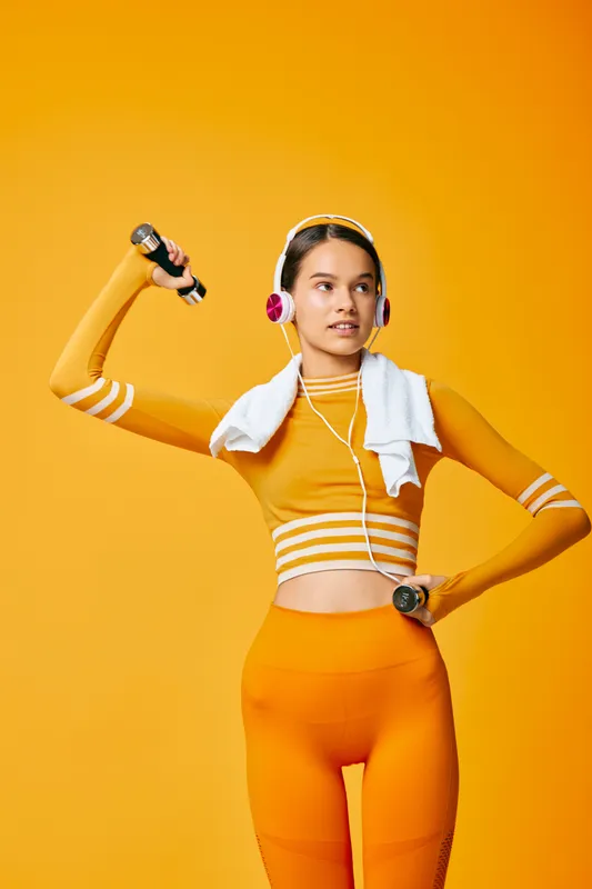 Energetic woman fitness athlete in orange outfit lifting a dumbbell, headphones on, towel draped, ready for workout in a vibrant studio with bold orange backdrop.