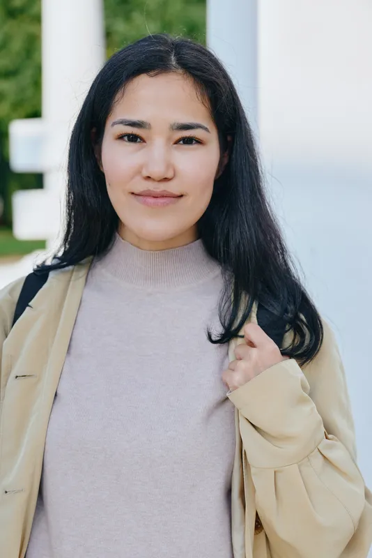 Woman, female, outdoors, casual fashion, smiling while wearing a light jacket and bag, showcasing a relaxed everyday style and approachable demeanor in a sunny setting.