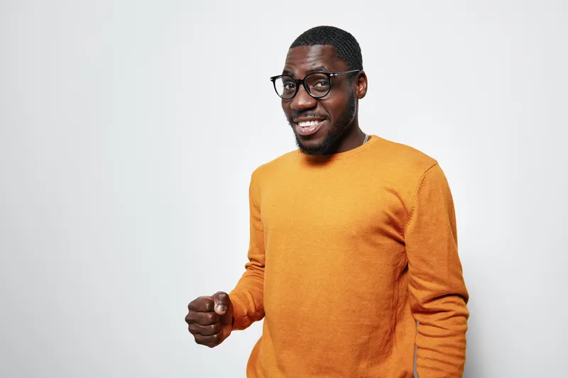 African American man with dark skin wearing glasses and orange sweater smiling confidently on white background. Portrait of happy black male with positive expression.