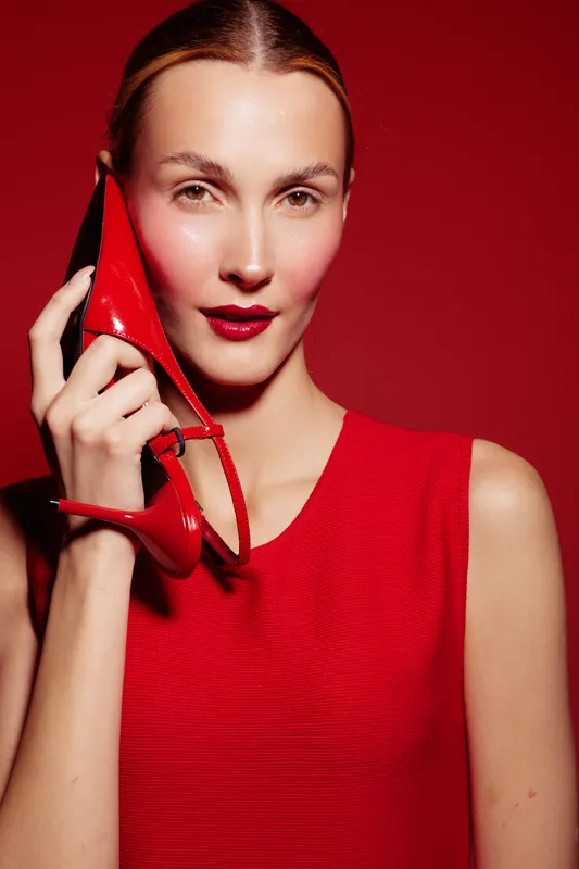 Young woman in red dress holds red high heel shoe near face, posing on red background. Concept of fashion, style, elegance, and beauty with vibrant color contrast.