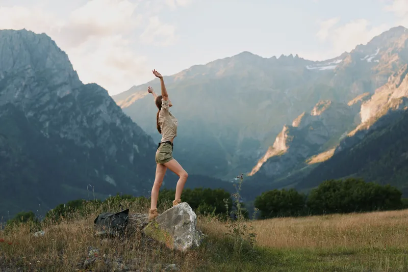 Young woman in casual clothes stands on a rock with arms raised in a mountain field, enjoying freedom and nature during a sunny day.
