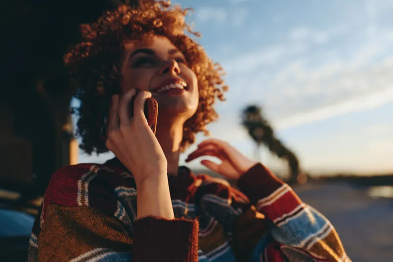 Woman with curly hair smiles while holding a smartphone to her ear, enjoying outdoors lifestyle at sunset, warm evening light, casual street background, candid moment of call, relaxed mood