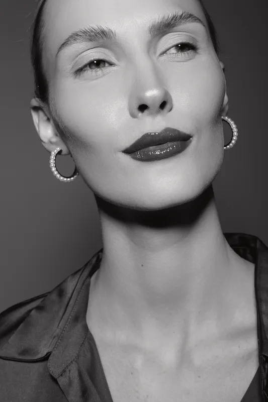 Close-up portrait of a fair skinned white woman with slicked back hair and glossy lips wearing hoop earrings. Studio lighting highlights her confident expression and smooth skin.