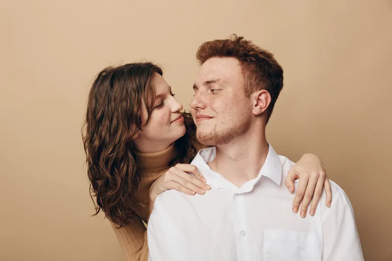 Young couple in casual clothes looks at each other with affection, woman embraces man from behind. Intimate moment, love, relationship, and happiness concept on beige background.