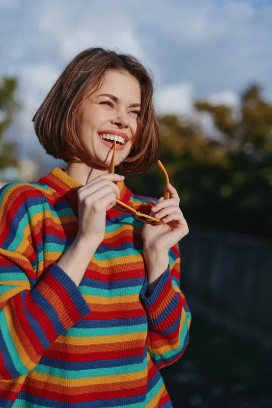 Woman smile sunglasses rainbow sweater portrait outdoors happy young female laughing while holding sunglasses near mouth, colorful knit jumper in natural light, candid joyful lifestyle image