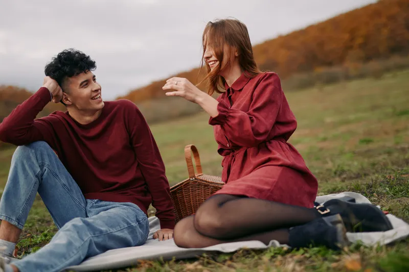 A young couple sharing a relaxed outdoor picnic, laughing and interacting with natural gestures that convey authenticity and credibility, captured in candid warm autumn light.