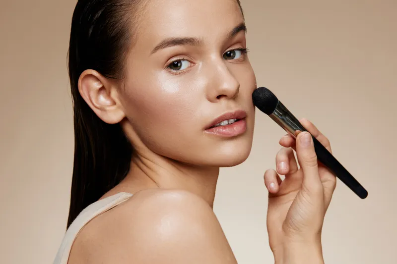 Young woman applying makeup with a brush, showcasing natural beauty, soft tone, and elegant skin Ideal for beauty and skincare concepts, including makeup tutorials