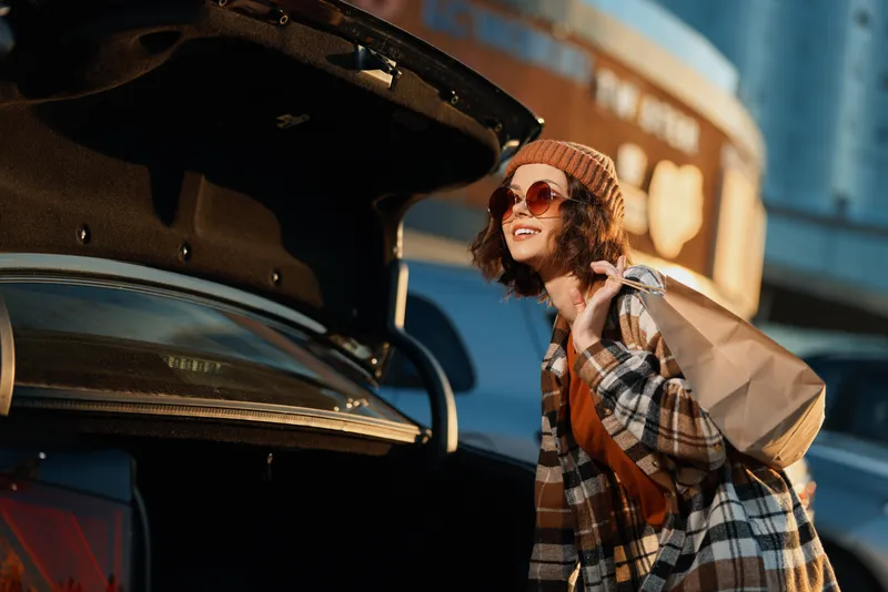 Woman by car trunk with bag and sunglasses wearing beanie and plaid coat, golden hour candid lifestyle portrait capturing authenticity, mindful living and emotional storytelling glow.