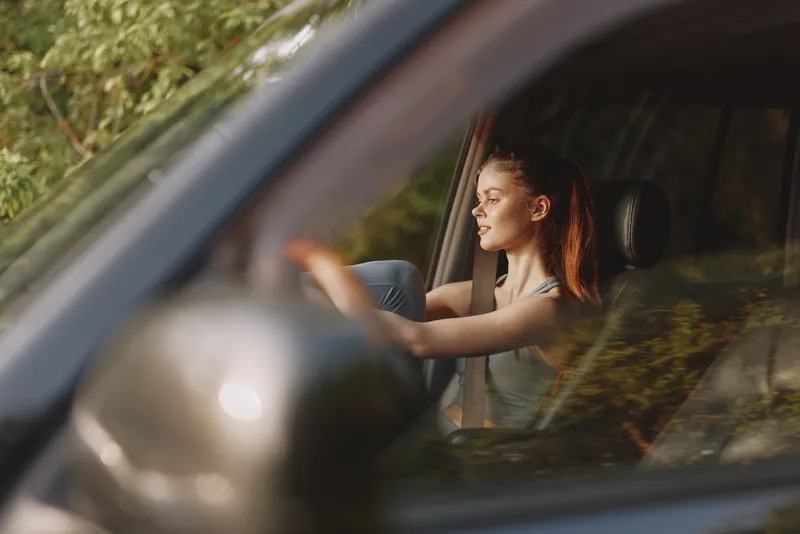 Young woman driving a car with a relaxed expression, enjoying nature and freedom, wearing casual clothing on a sunny day