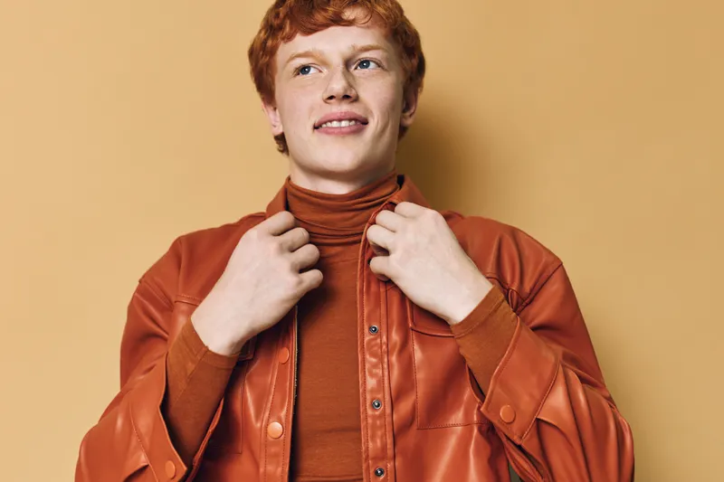 Young man with red hair wearing orange leather jacket and turtleneck looking away with smile on beige background. Portrait, fashion and casual style with confident expression and modern look.